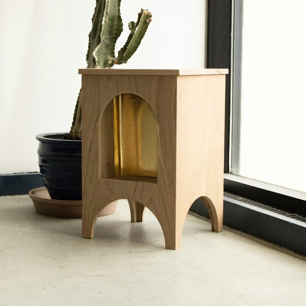 Mesa auxiliar de roble con un hueco en forma de arco y patas recortadas, junto a un ventanal y una maceta con cactus