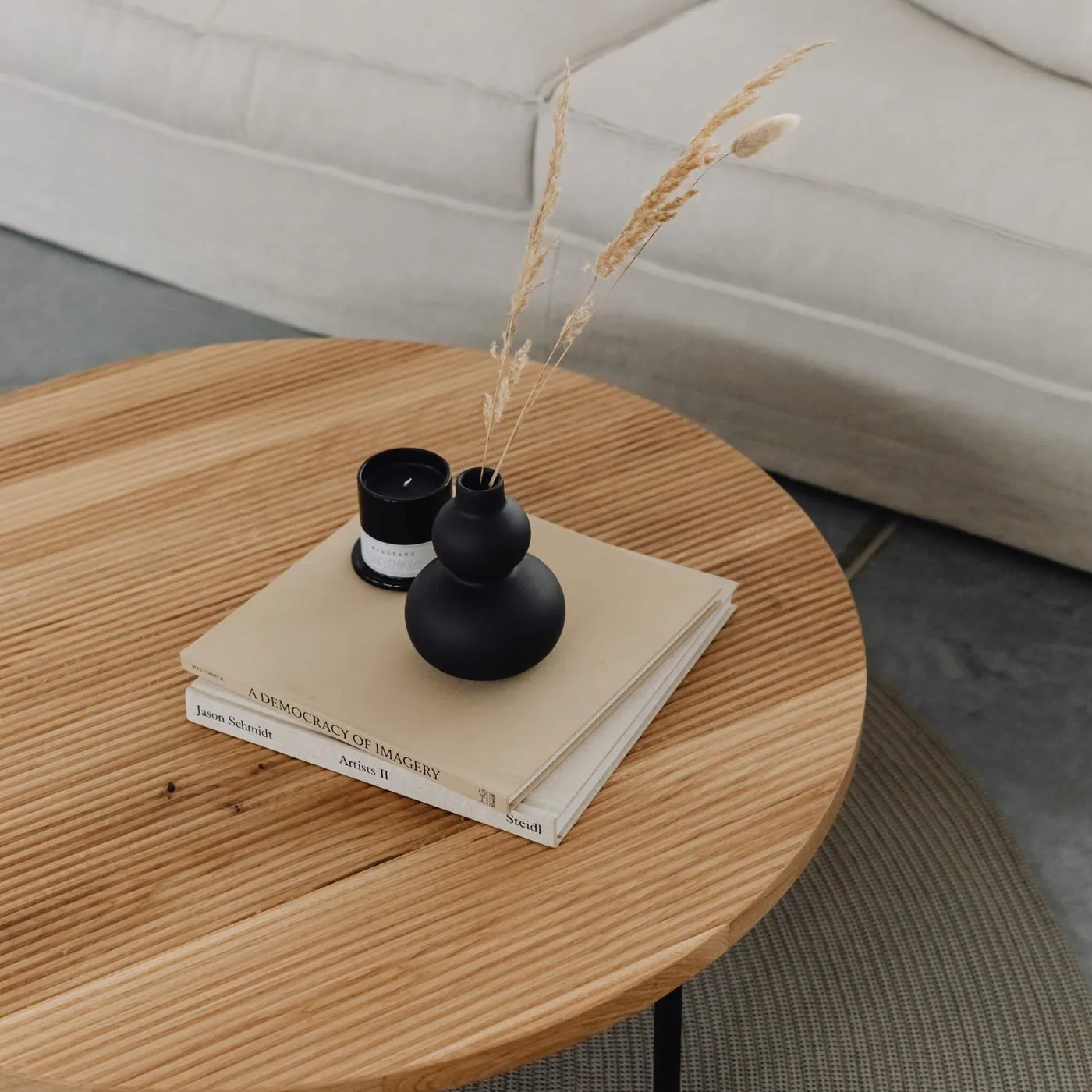 Mesa ratona redonda de roble con libros, una vela y un florero negro, frente a un sillón claro